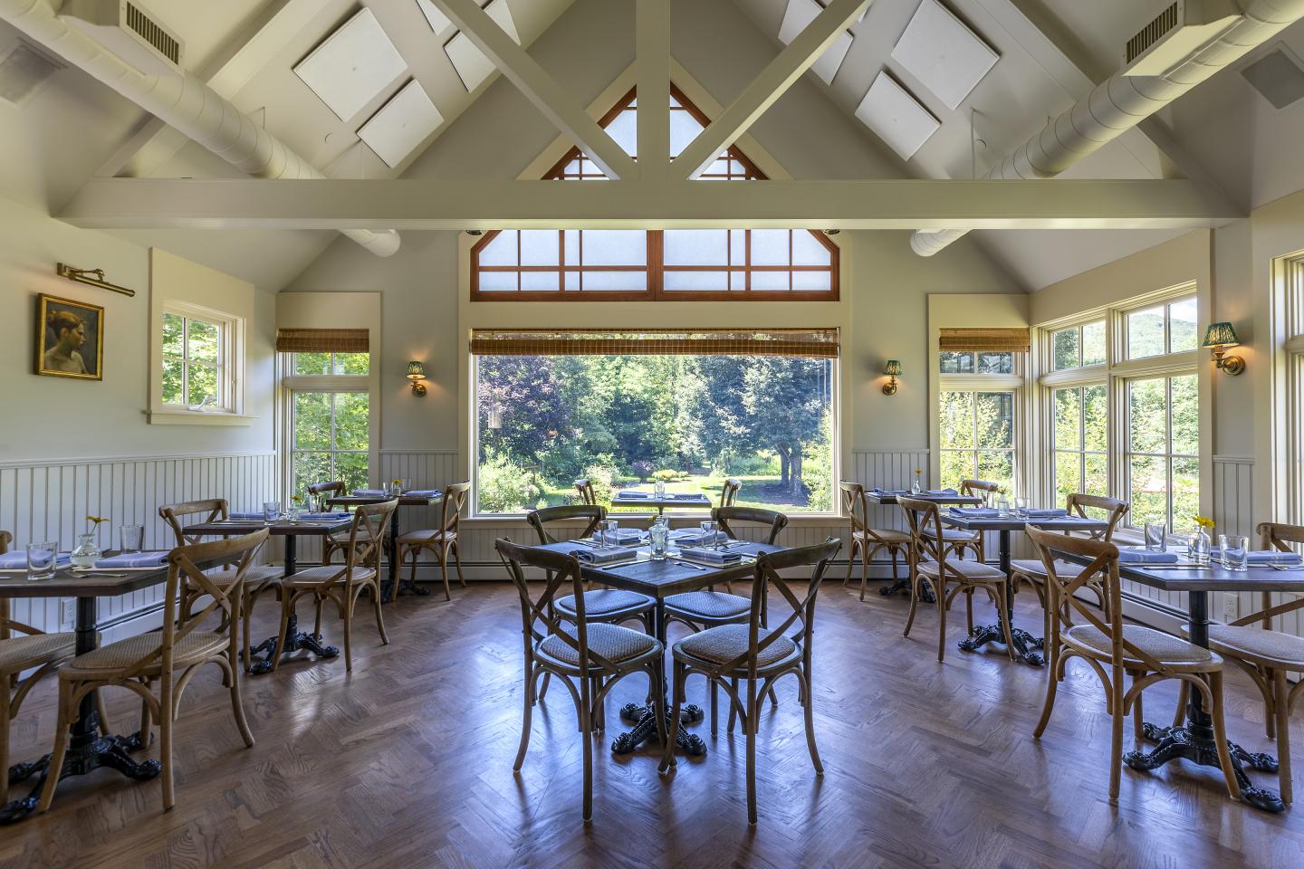 Bright restaurant interior with wooden tables, chairs, and large windows facing a garden.