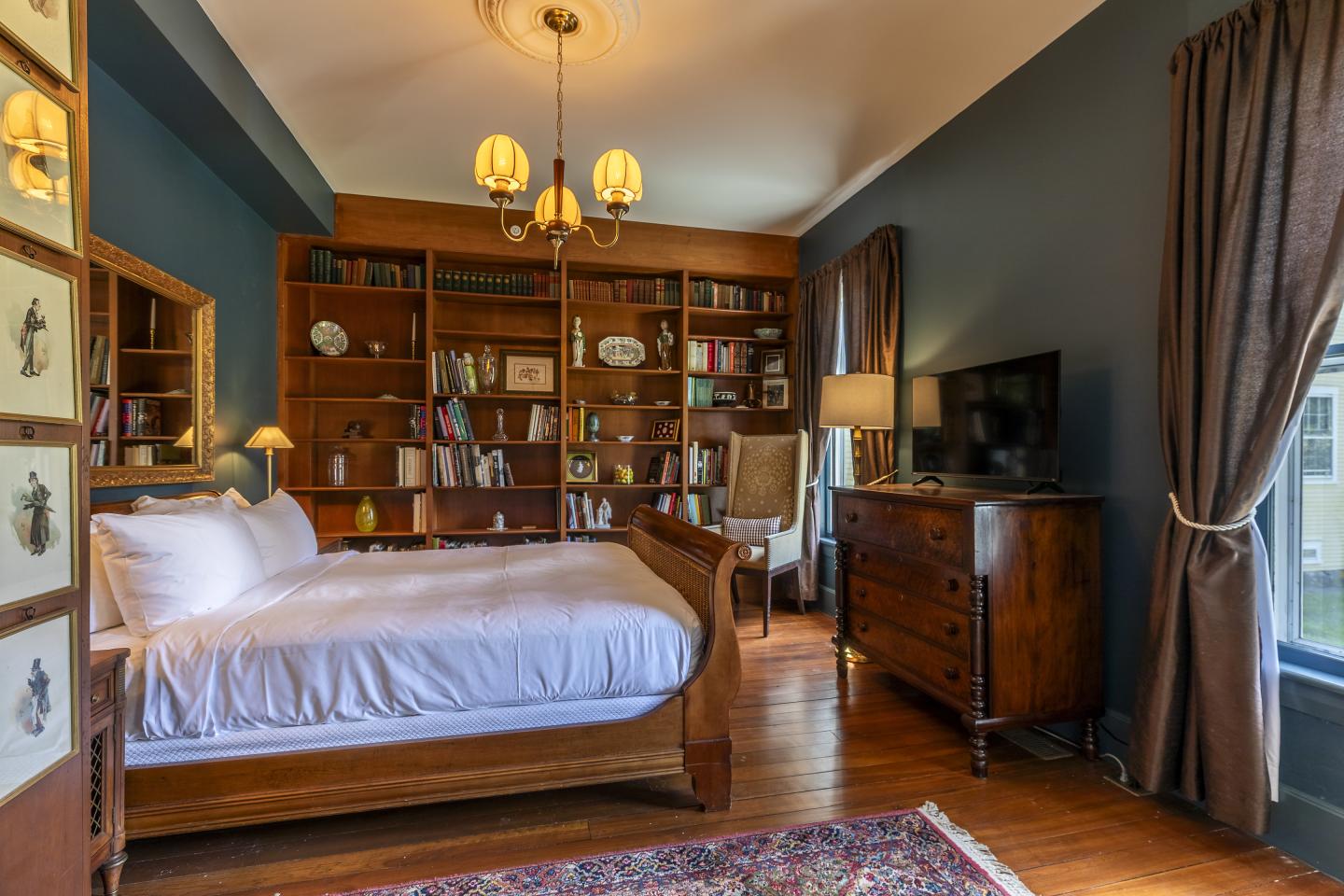 Elegant bedroom with wooden furniture and bookshelves, warm lighting, and dark curtains.