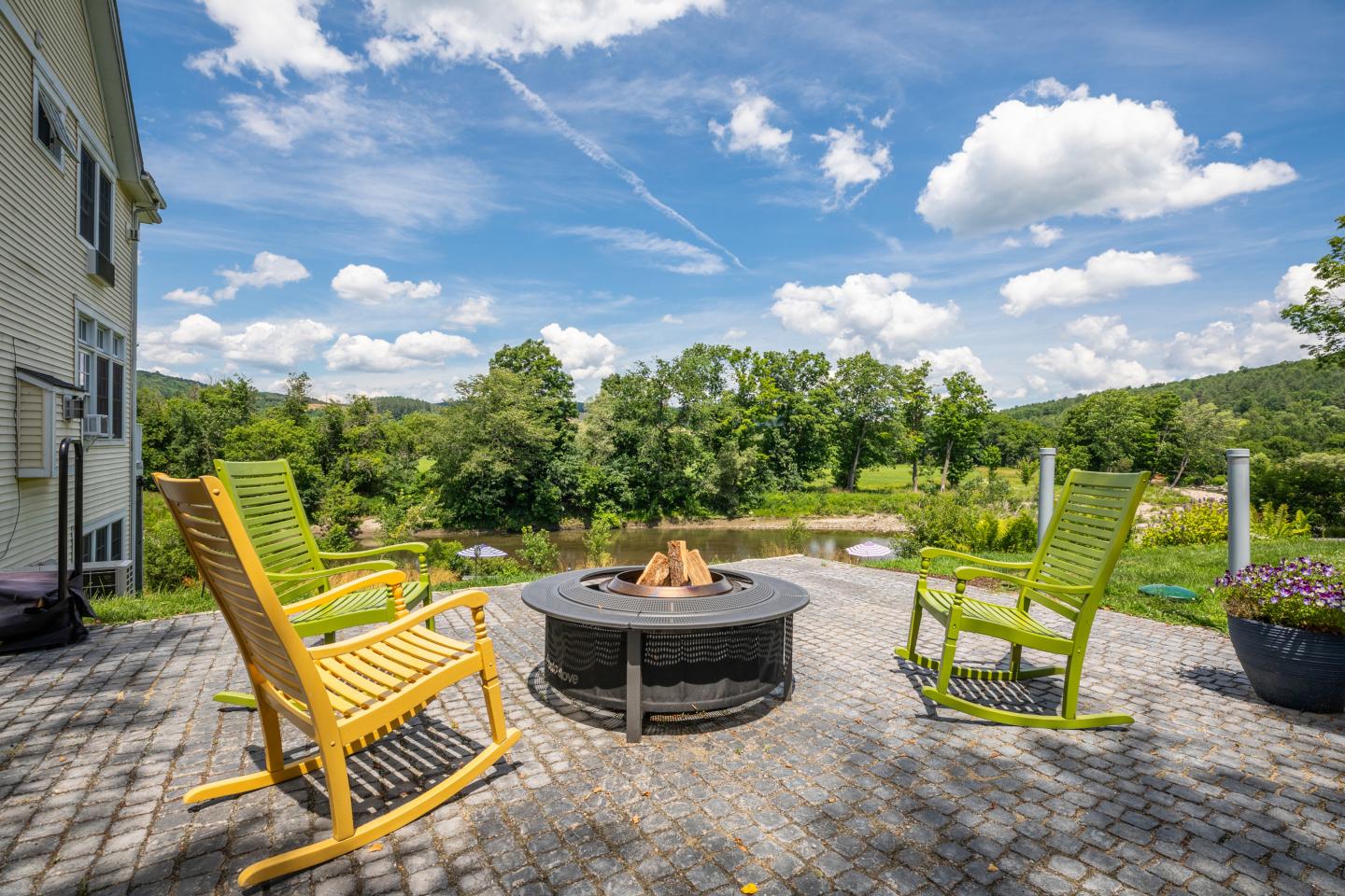 Sunny patio with three chairs around a fire pit, overlooking river, green hills and trees.