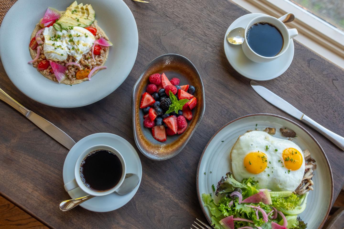 Breakfast dishes with eggs, berries, and two cups of black coffee on a wooden table.
