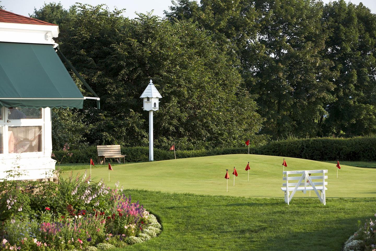 Sunny golf course with putting green, red flags, trees, and white bench.