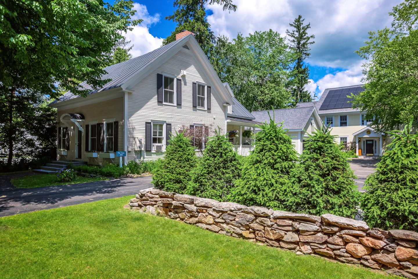 Cape house with green lawn, stone wall, and tall trees under a blue sky with clouds.