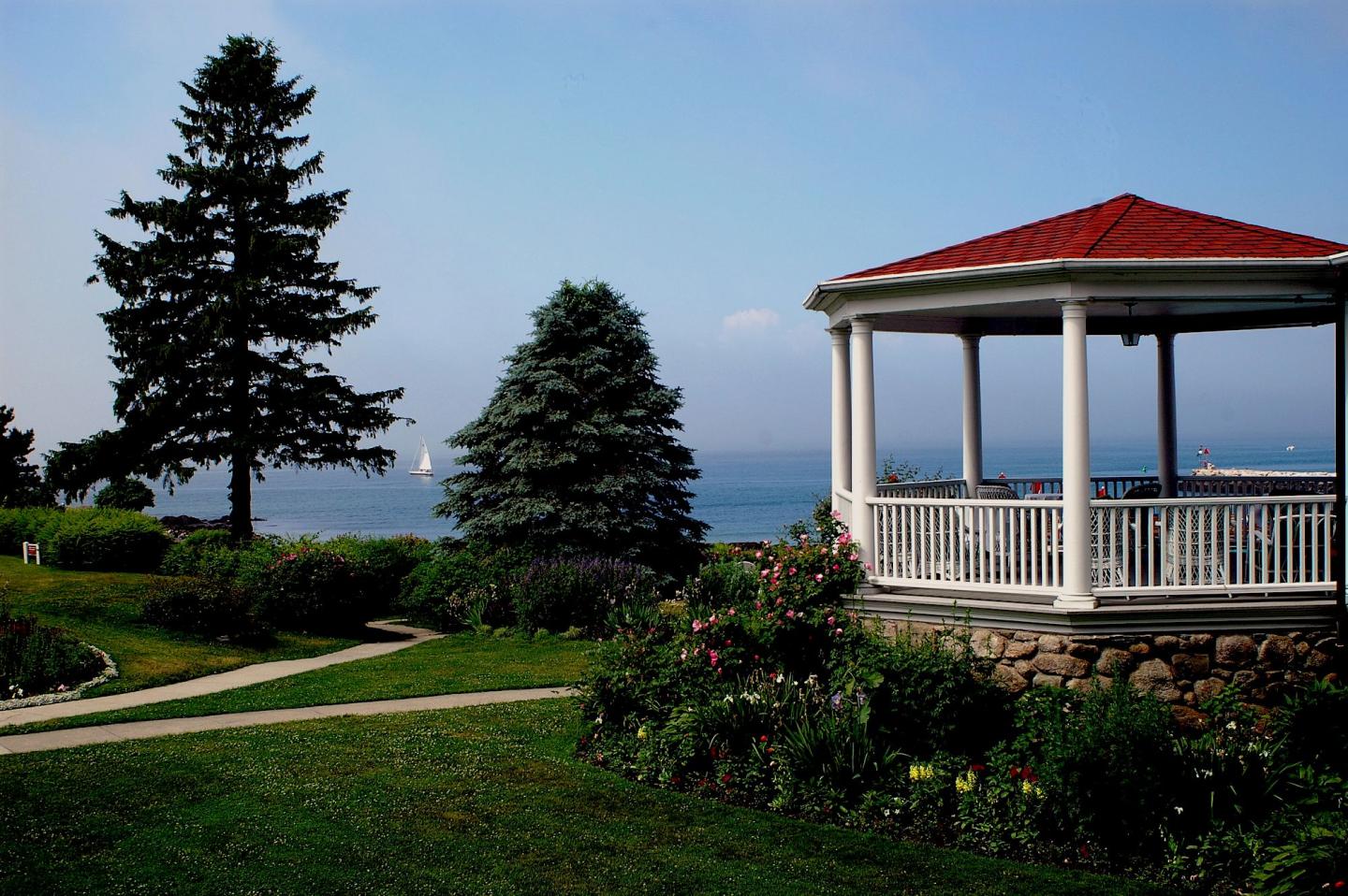 Gazebo with red roof by the sea, surrounded by lush greenery and two large trees.
