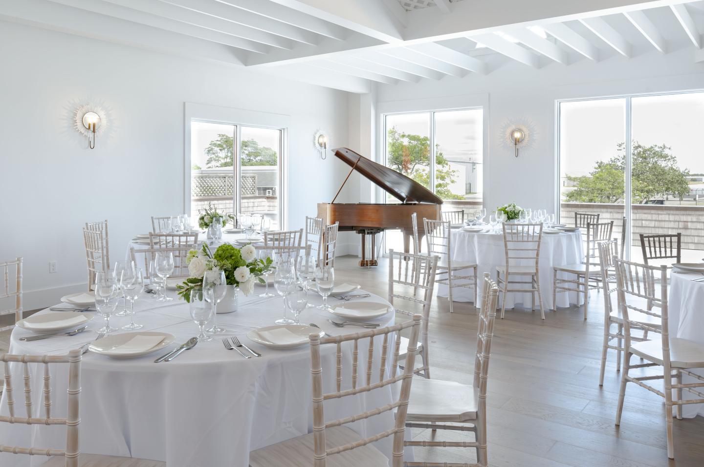 Bright banquet room with round tables, white decor, and a grand piano by the window.