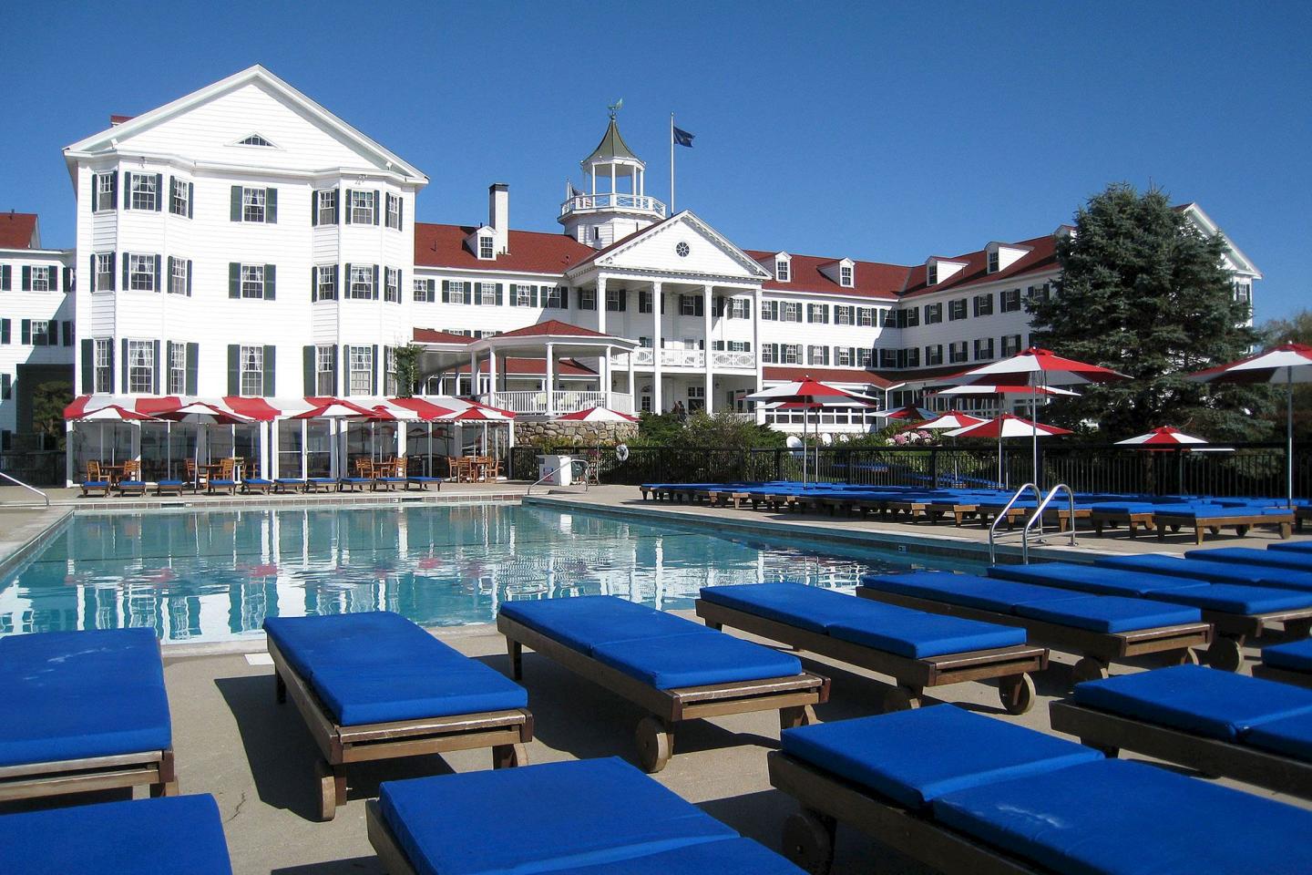 White hotel with red roofs, pool, and blue lounge chairs under a clear sky.
