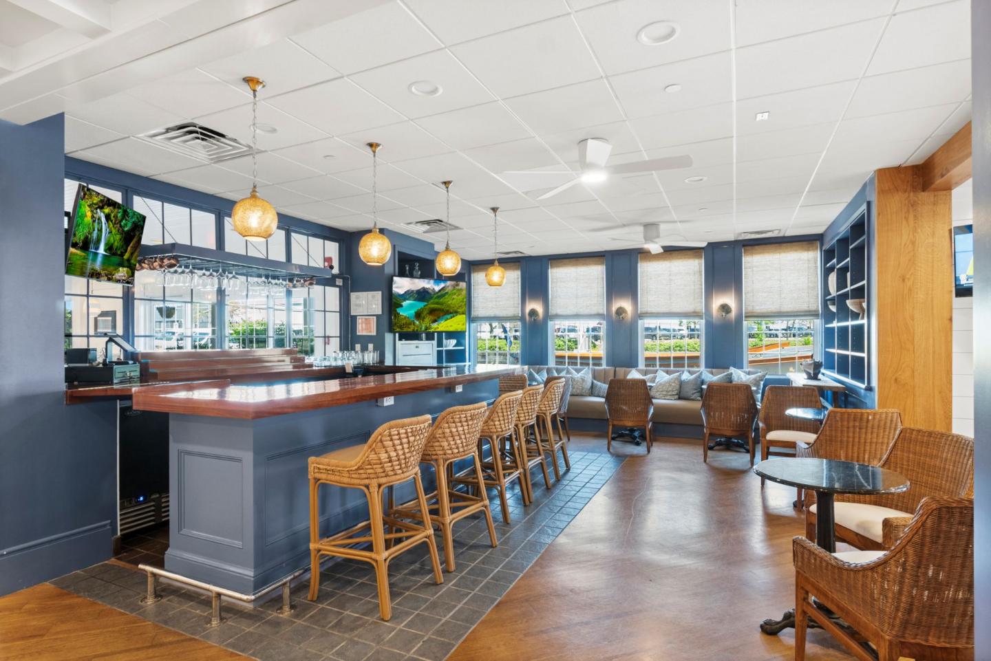 Bar with wooden stools, tables, blue accents, and TVs on walls. Bright with natural light.
