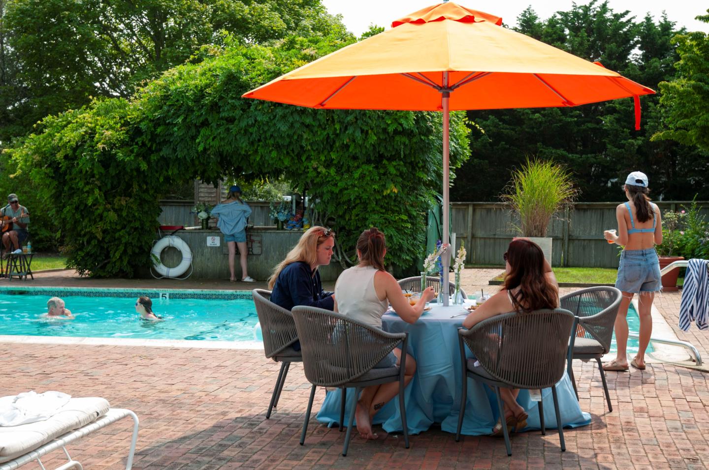 Poolside table with orange umbrella, three people seated, others swimming in the background.