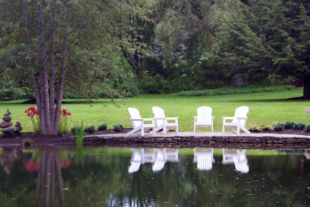 Four white chairs on a grassy shore facing a calm pond, surrounded by trees.