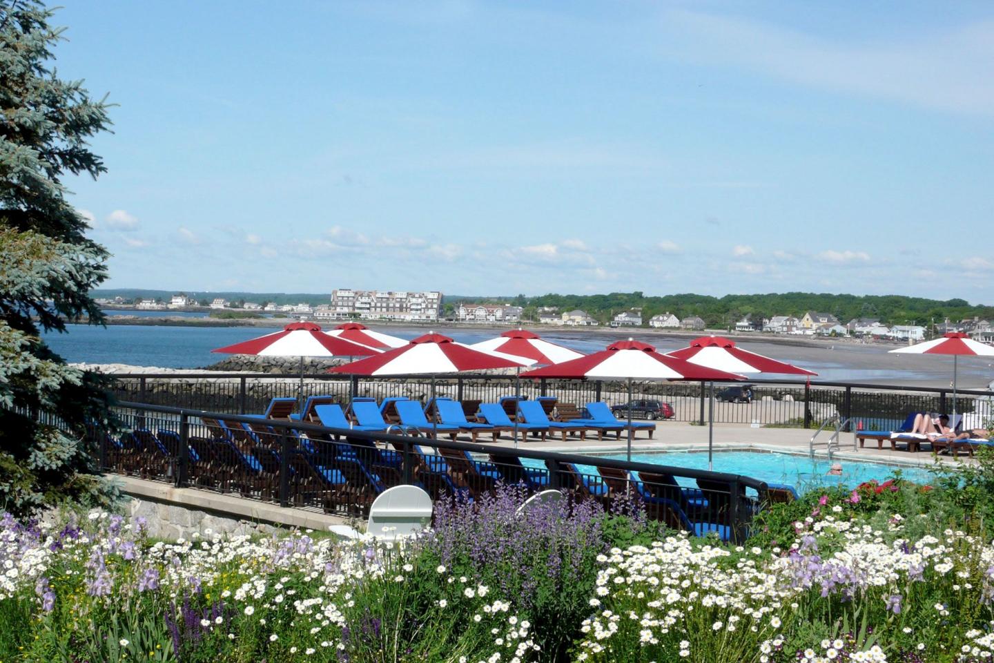 Poolside loungers with red umbrellas, overlooking a calm sea and distant town.