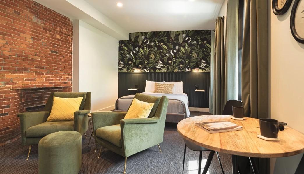 Cozy hotel room with green chairs, bed, and table. Brick and botanical accent walls.