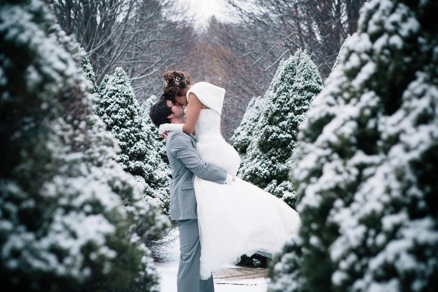 Bride and groom embrace in snowy forest, surrounded by evergreen trees.
