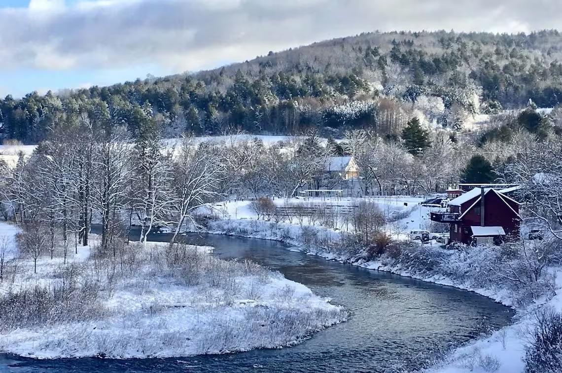 Snowy landscape with a river, red barn, and hills in the background.
