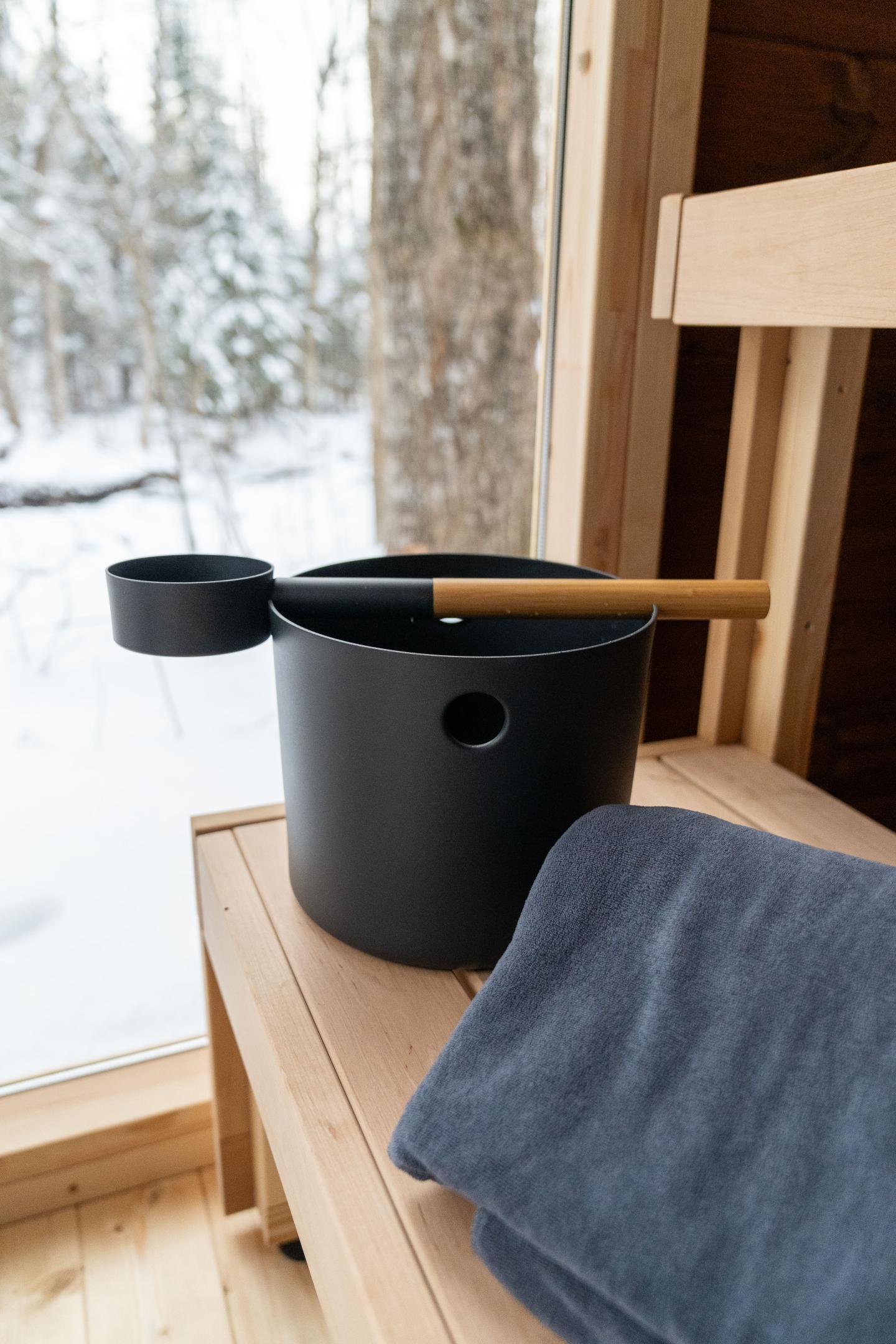 Sauna bucket and ladle on a wooden bench with a snowy view outside.