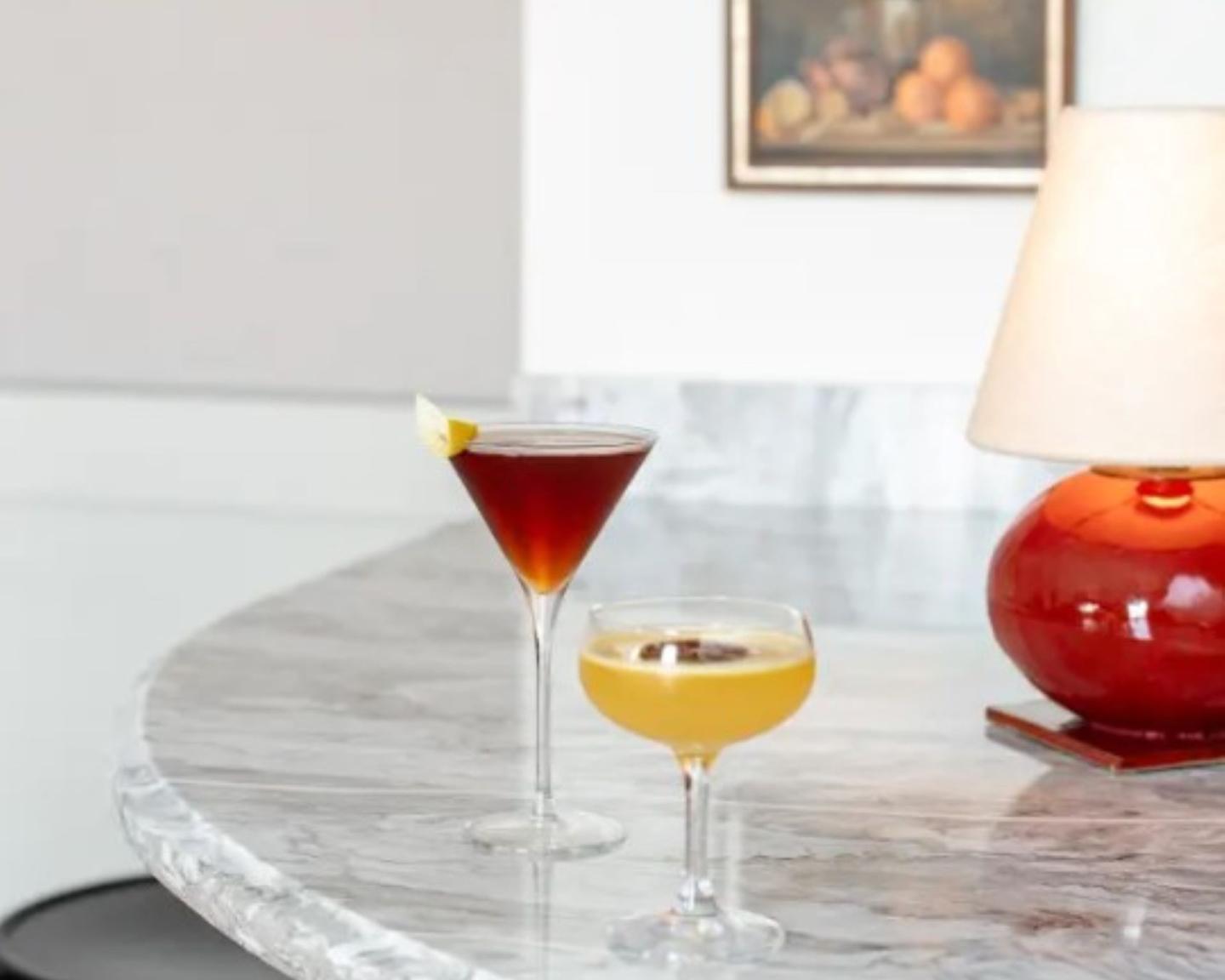 Two cocktails on a marble counter, one red, one yellow, beside a red lamp.
