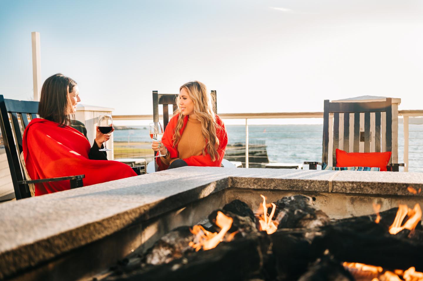 Two women relax by a seaside fire pit, wrapped in red blankets.