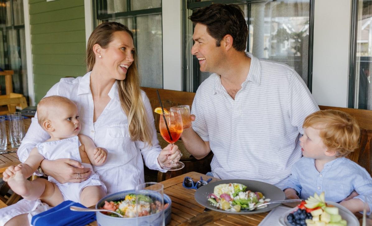Family dining outdoors, smiling at each other, with food and drinks on the table.