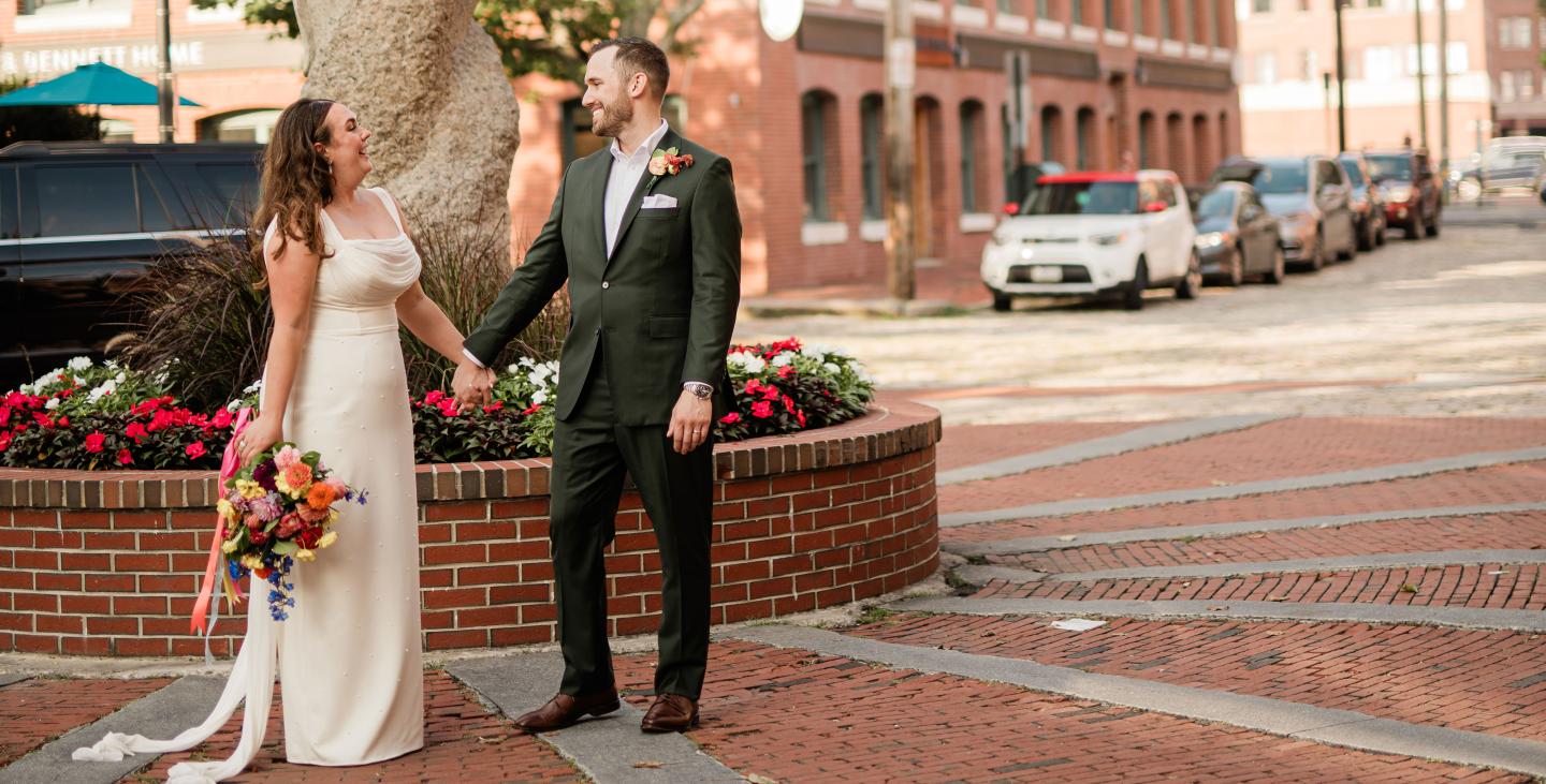 Bride and groom smiling, holding hands on a street with flowers and parked cars.