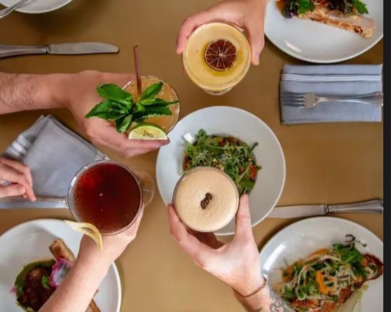 Four hands toasting with cocktails over plates of food on a table.