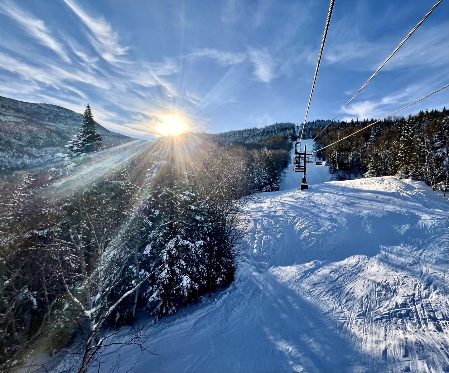 Snowy mountain landscape with a ski lift under a bright sun and blue sky.