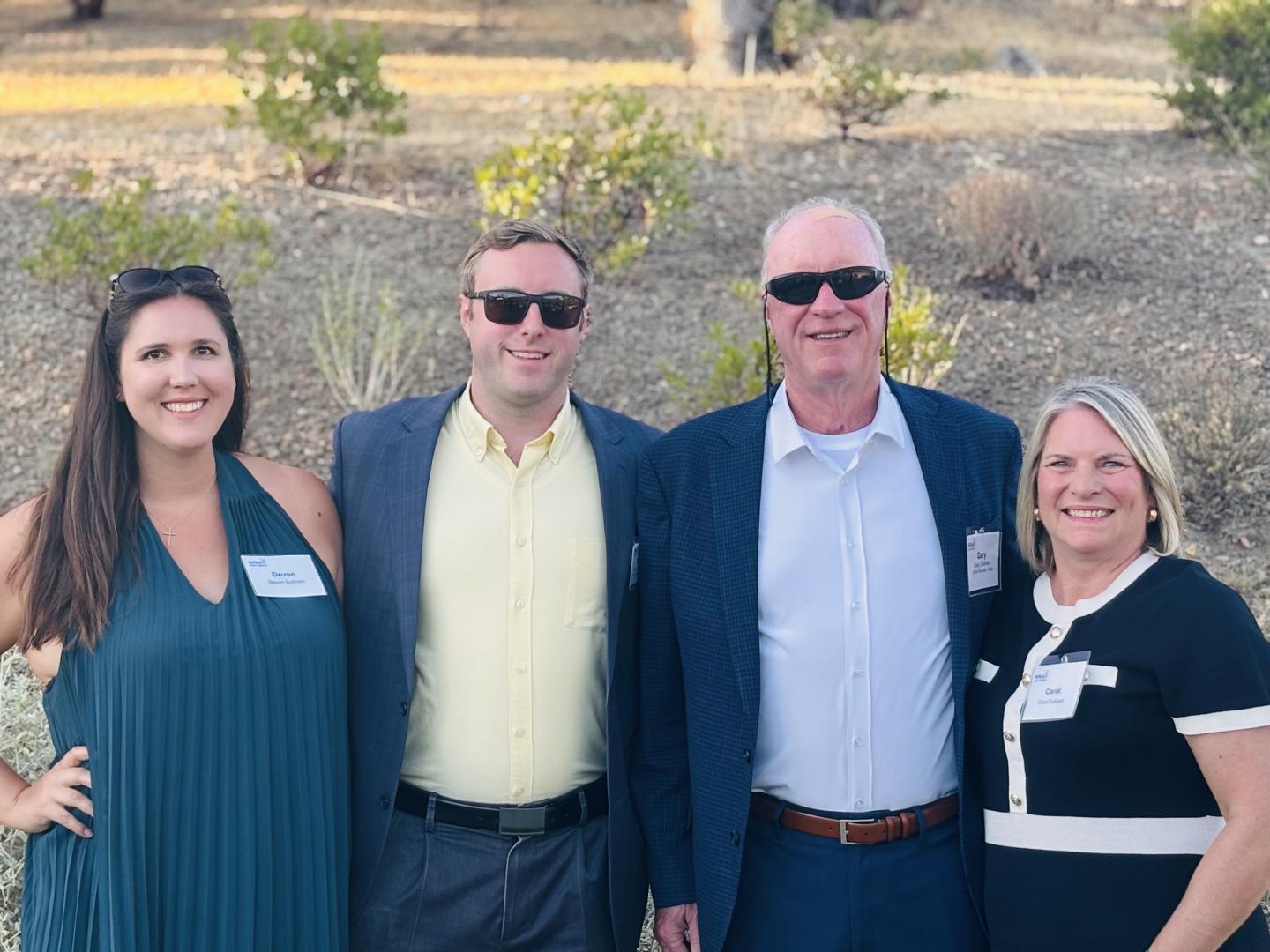 Four smiling people outdoors, all wearing name tags and sunglasses.