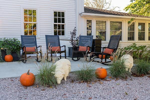 Black rocking chairs with orange cushions on porch, pumpkins and bushes in front.