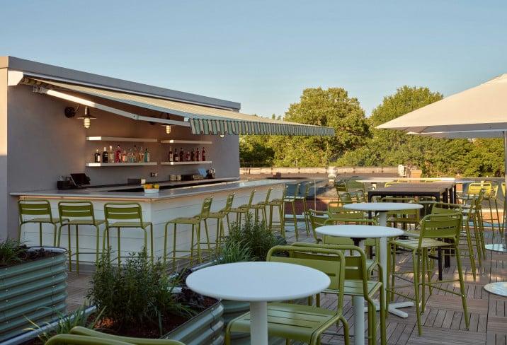 Rooftop bar with green chairs, white tables, and awning, surrounded by trees.