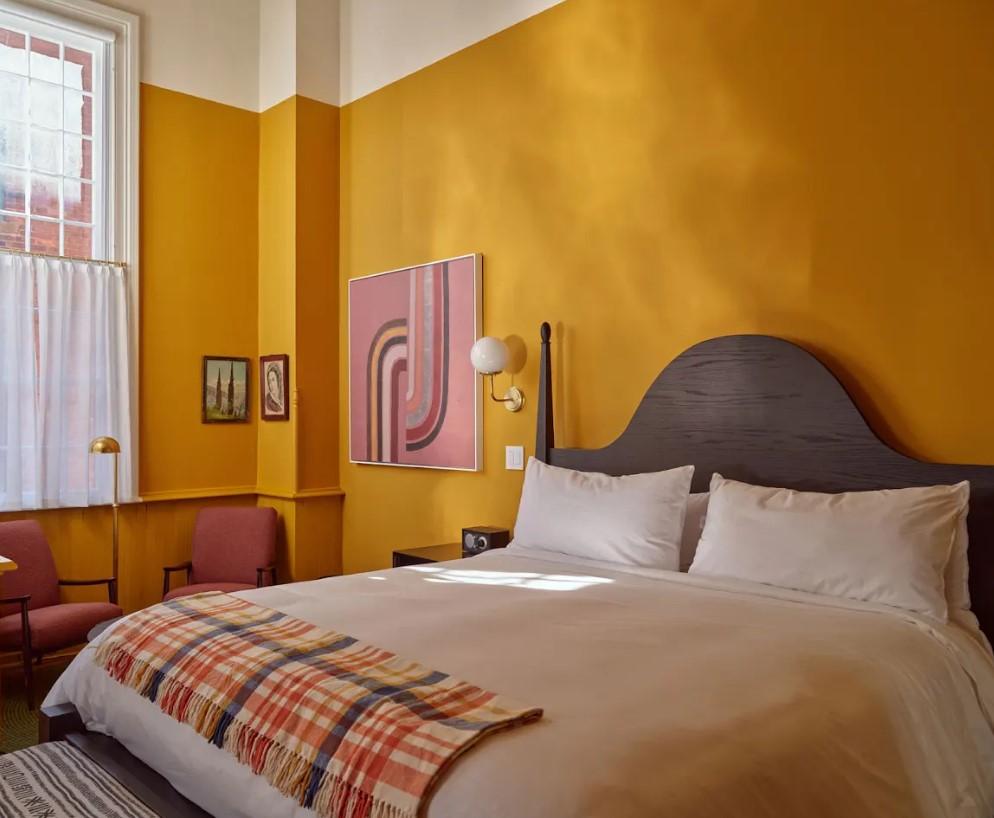 Cozy bedroom with mustard walls, white bedding, plaid blanket, and abstract art.