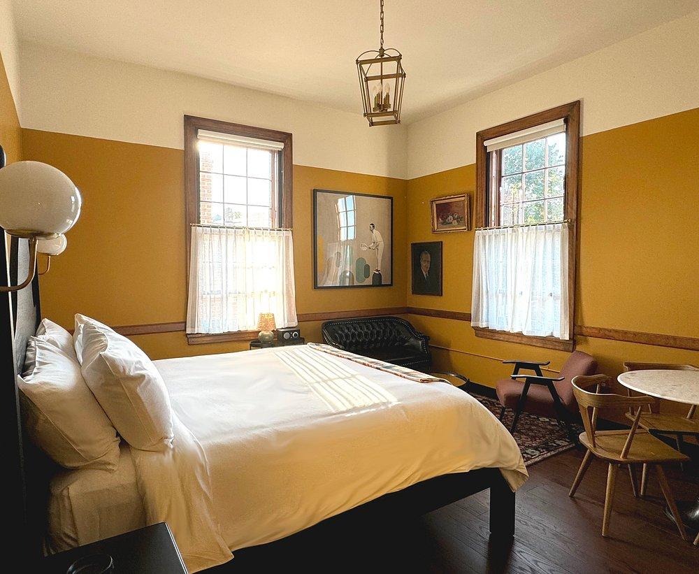 Cozy bedroom with yellow walls, a bed, two windows, and a small seating area.