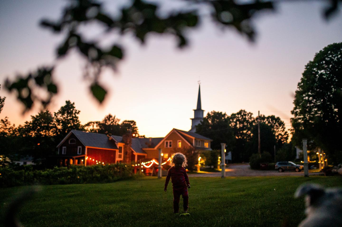 Child in a yard at dusk, facing lit-up buildings and a church steeple.