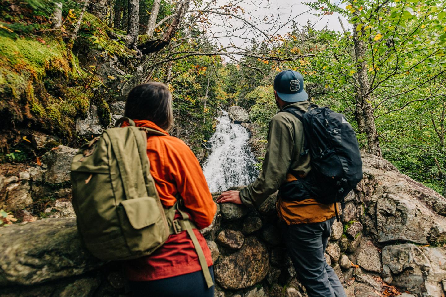 Two hikers with backpacks near a forest waterfall.