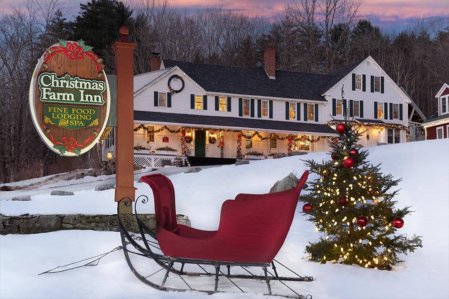 Red sleigh and Christmas tree in snow, lit inn with festive decor in the background.