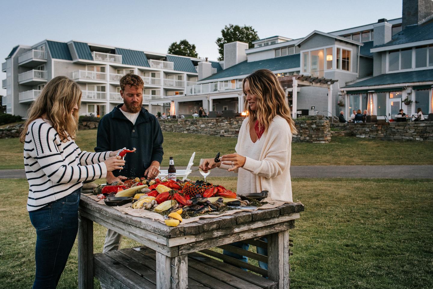 Three people preparing food on a table outside a coastal house.