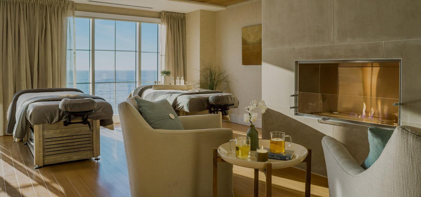 Spa room with two massage tables, comfortable chairs, and a fireplace. Ocean view through windows.