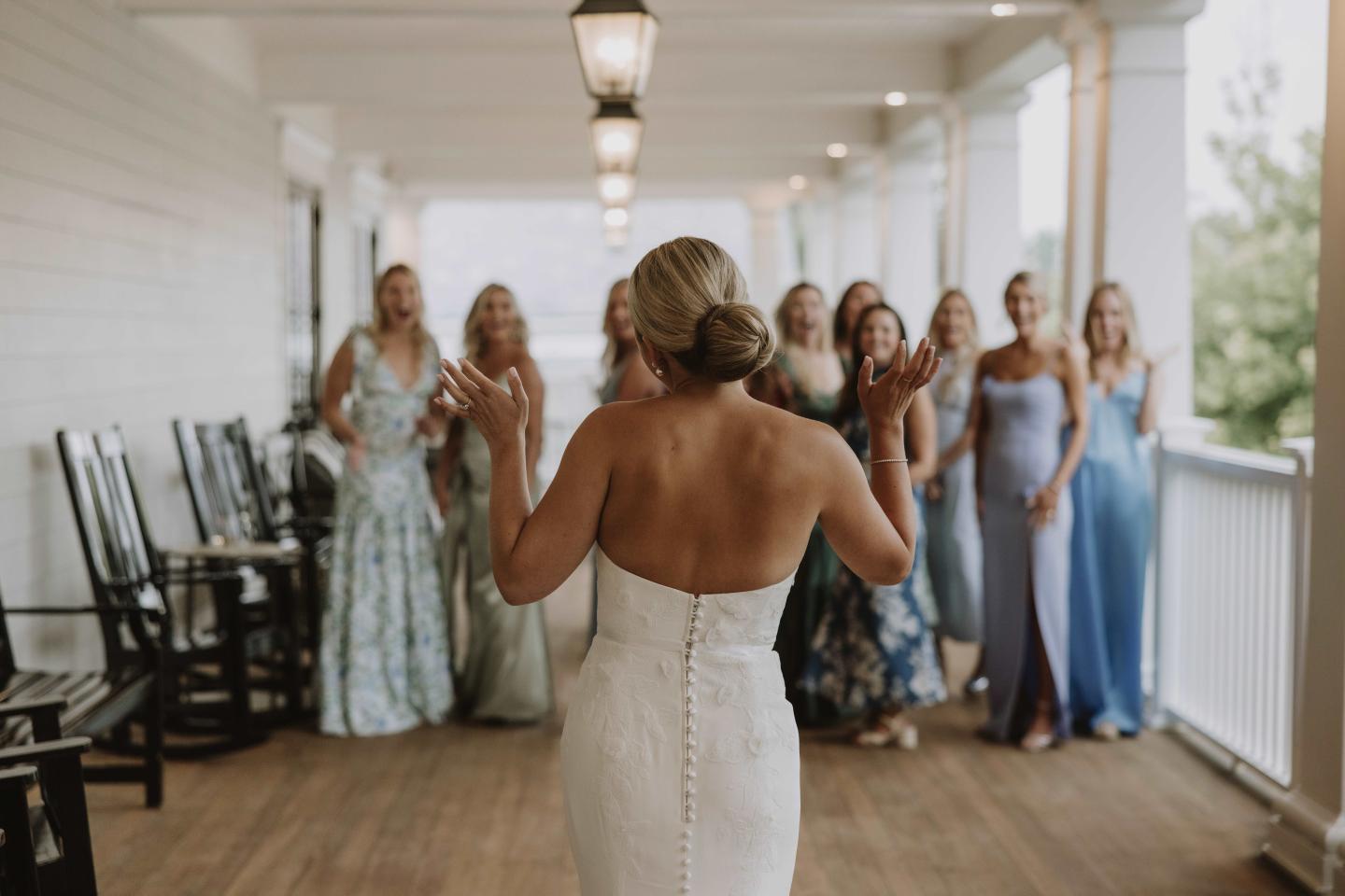 Bride in a white dress faces bridesmaids in colorful dresses on a porch.