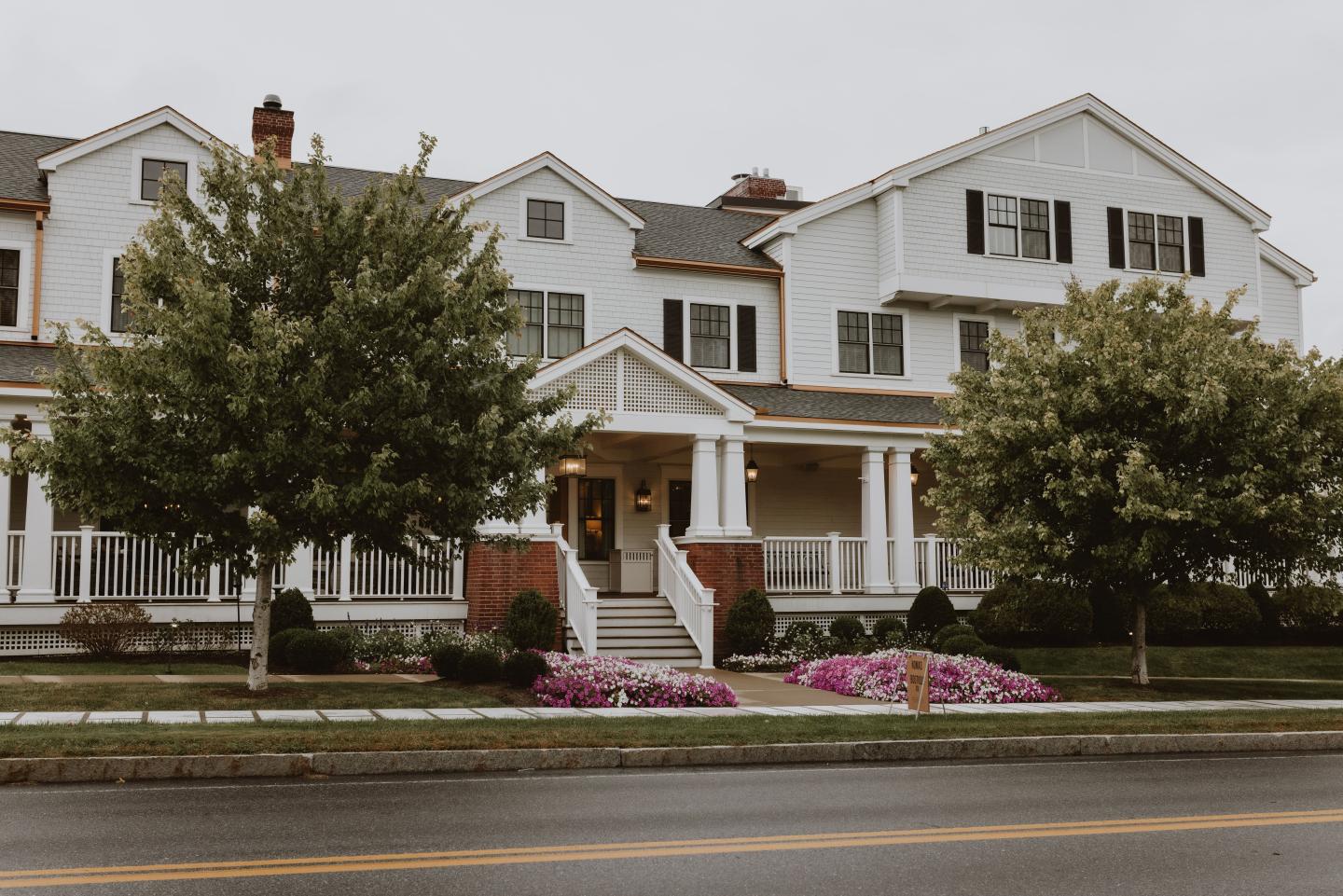 Large white house with a porch, flanked by trees and flowerbeds. Overcast sky.