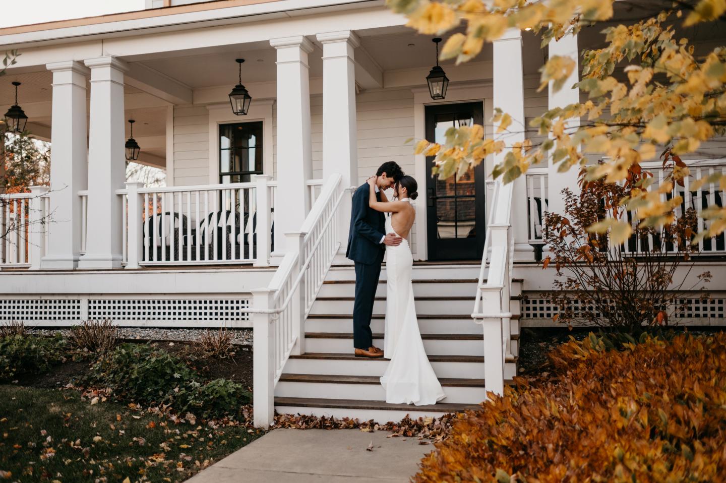 Bride and groom embrace on porch steps of a white house in autumn.