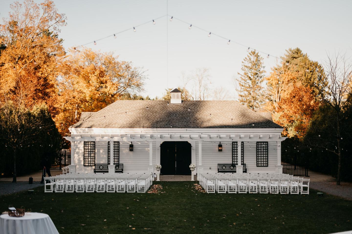 White house with autumn trees, string lights, and arranged chairs on grass.