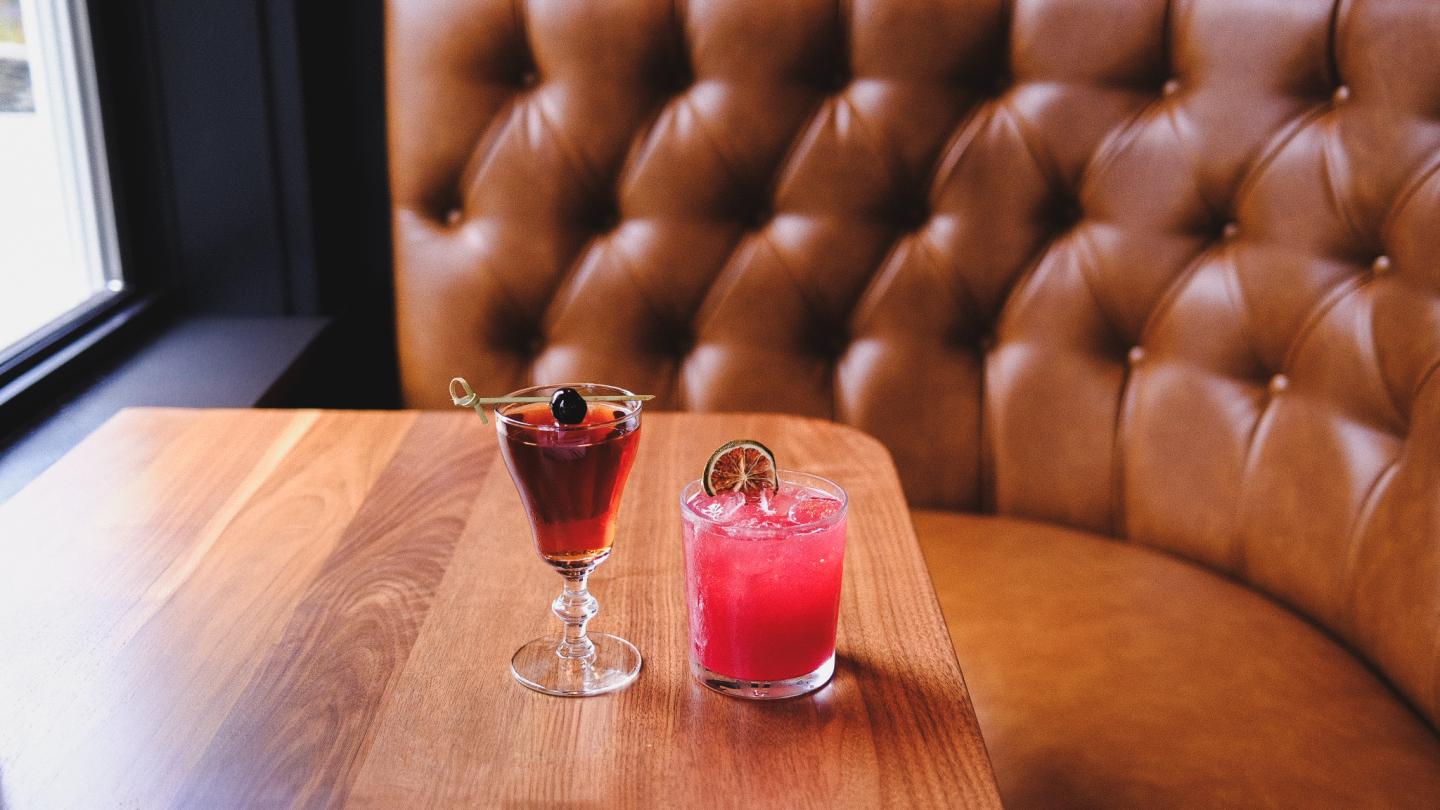 Two cocktails on a wooden table, with a leather booth background.