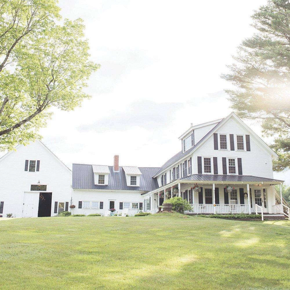 Large white farmhouse with porch, surrounded by green grass and trees in sunlight.