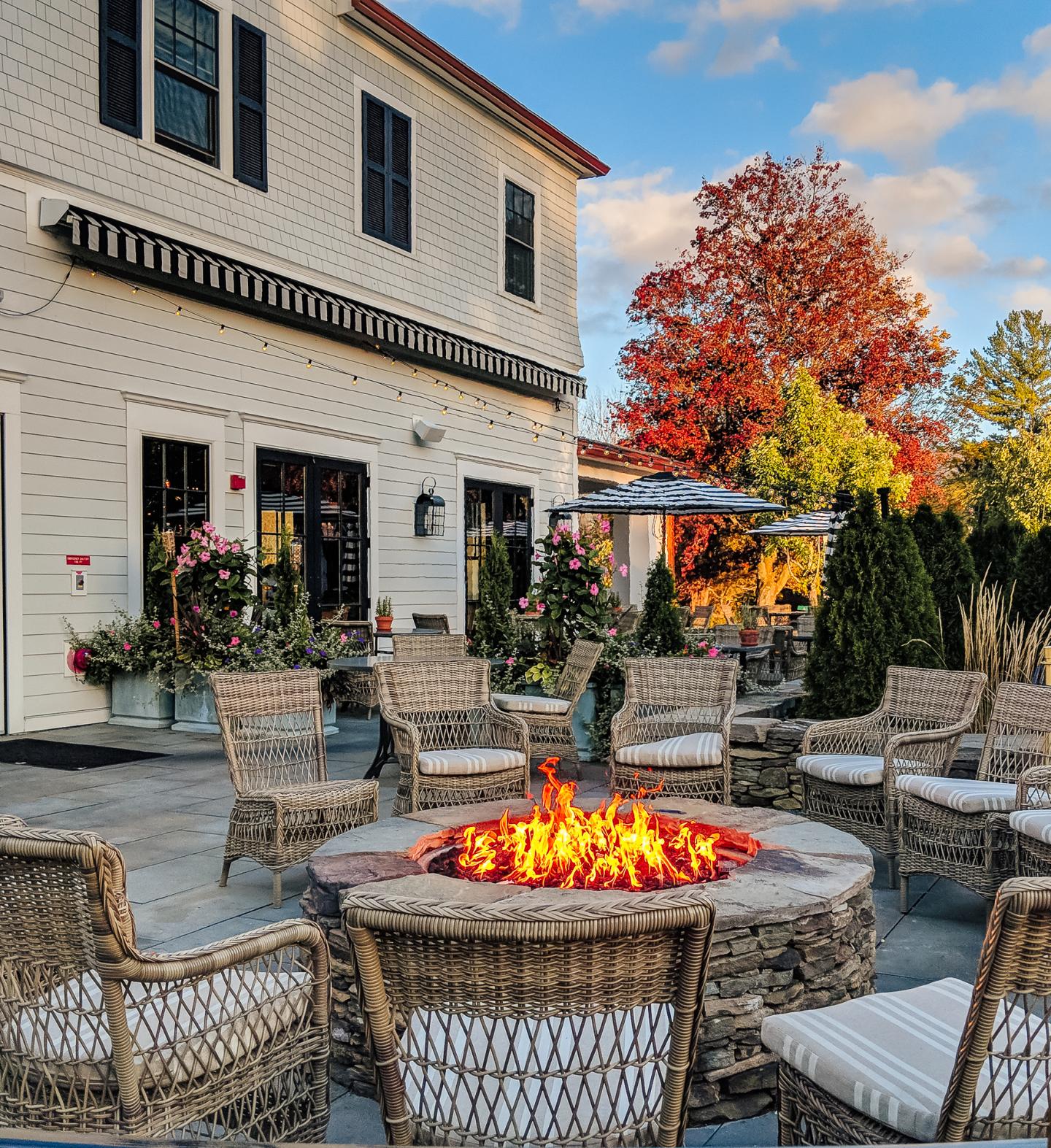 Fire pit surrounded by wicker chairs on patio, autumn trees in the background.