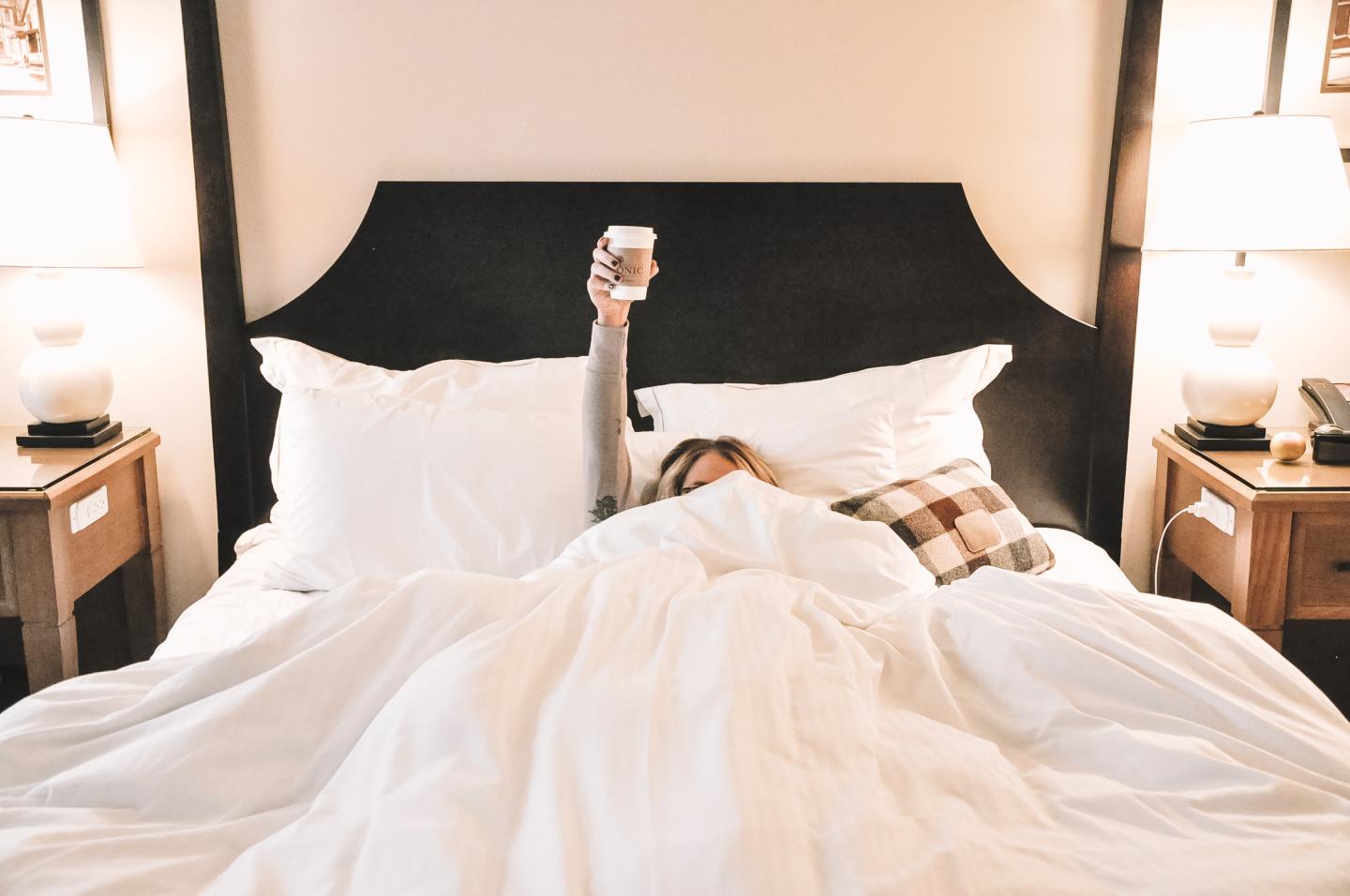 Cozy bed with person holding a coffee cup, white bedding, and nightstands with lamps.