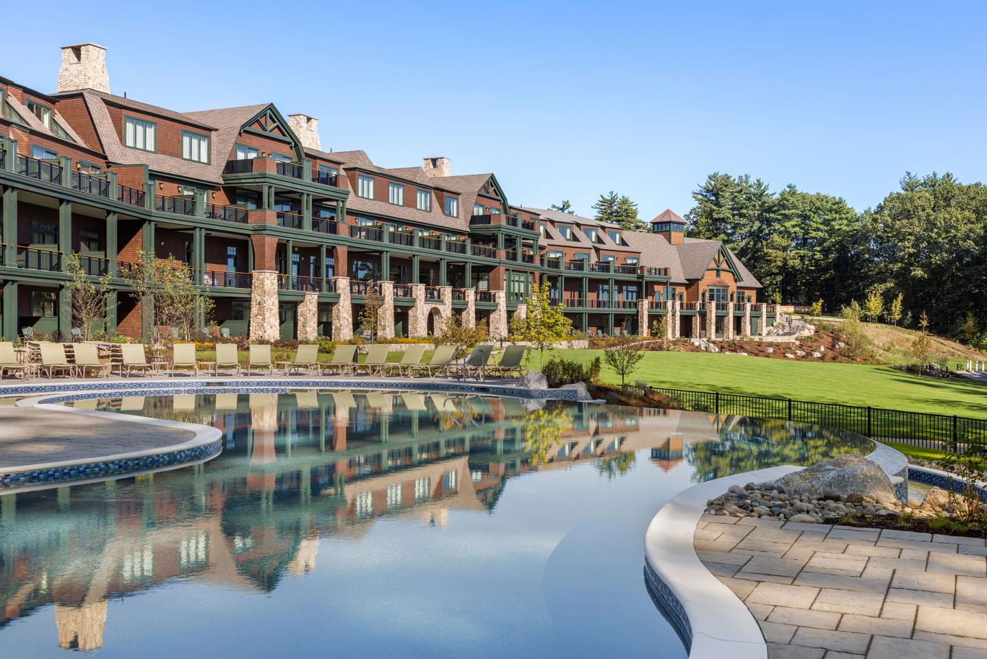 Large lodge with rustic design by a pool and green lawn under blue sky.