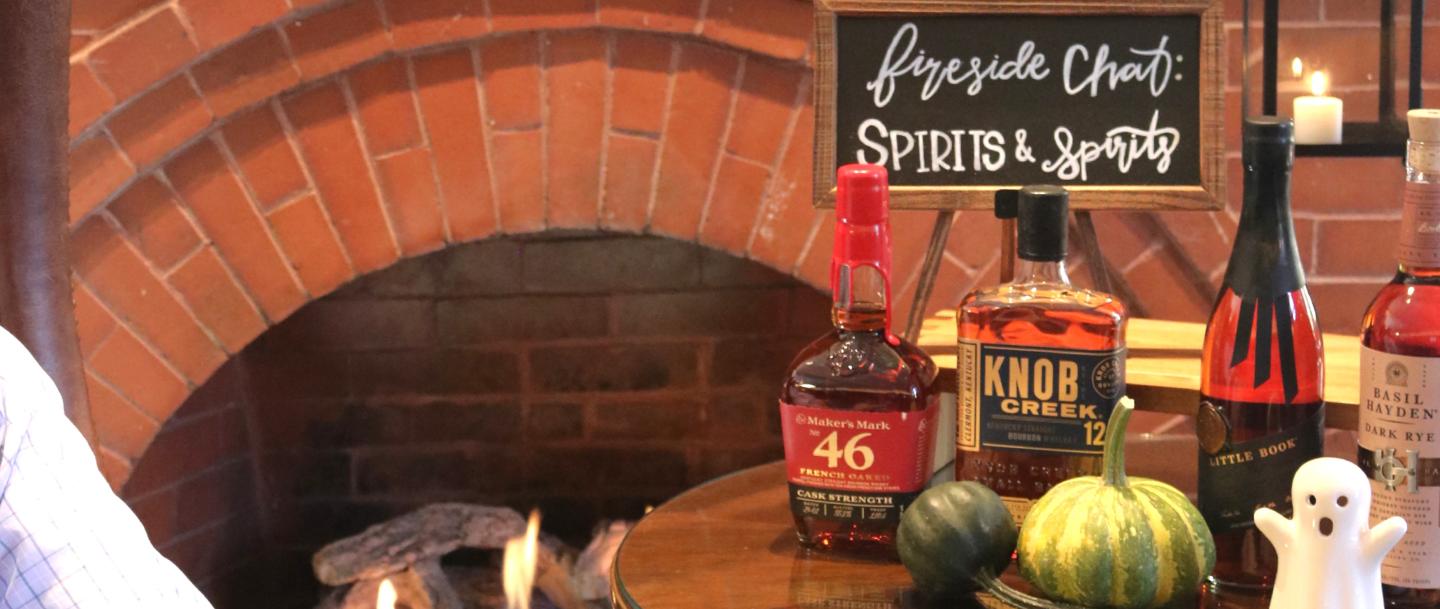 Bottles on a table with pumpkins and a ghost figure, against a brick arch backdrop.