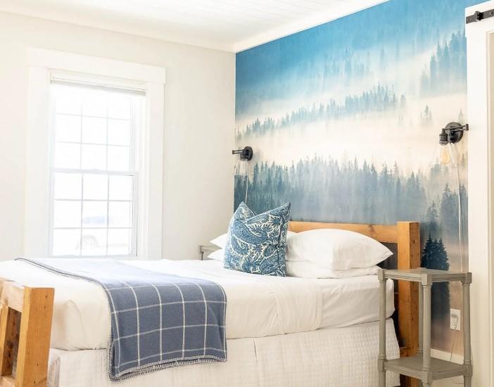Cozy bedroom with forest mural, white bed, blue pillows, and large window.