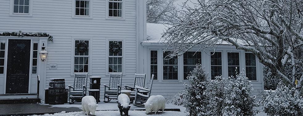 Snow-covered house with a porch, rocking chairs, and trees in a winter setting.