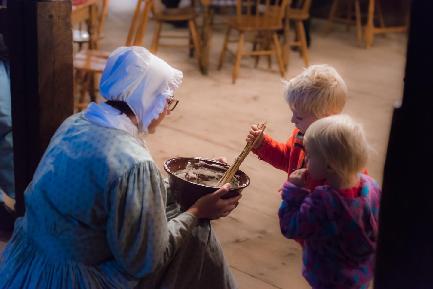 Woman in period dress shows two kids how to mix dough in a wooden room.
