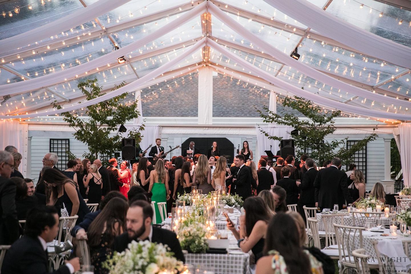 Wedding reception under a lit glass tent, guests mingling, band playing music.