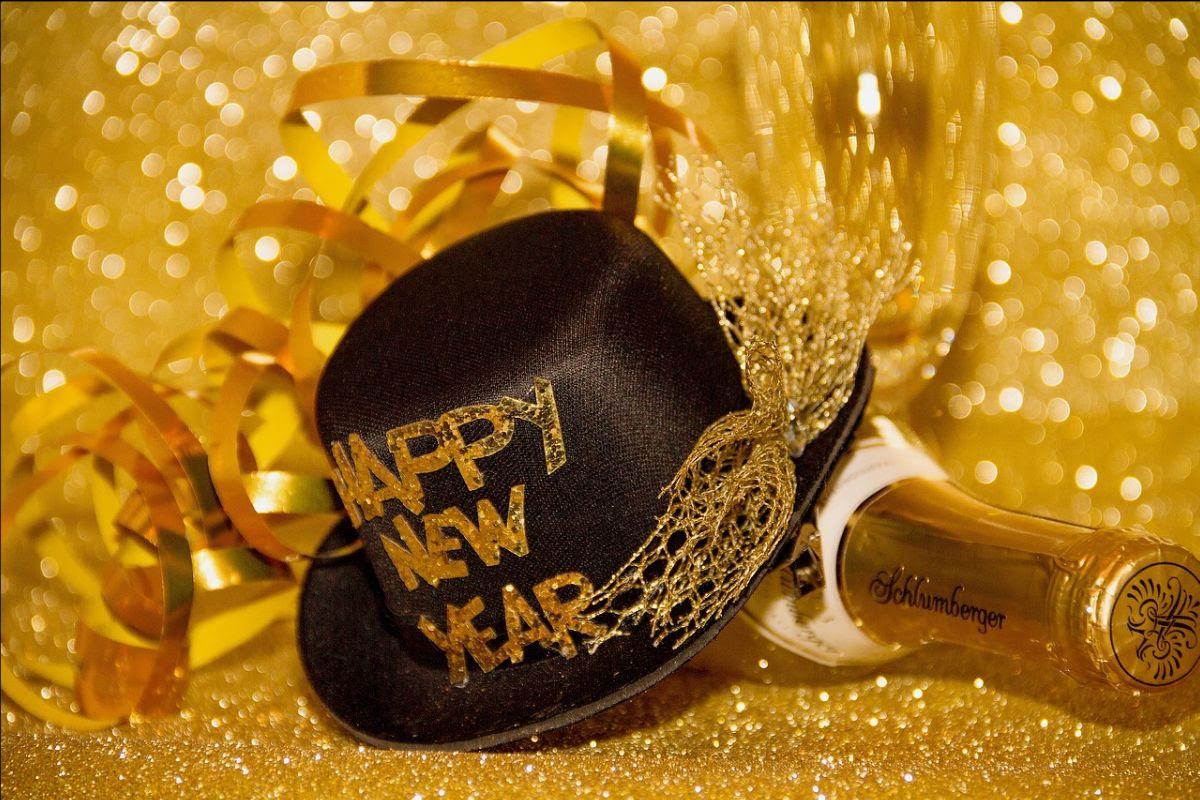 Black hat and champagne bottle with gold ribbons on a glittery gold background.