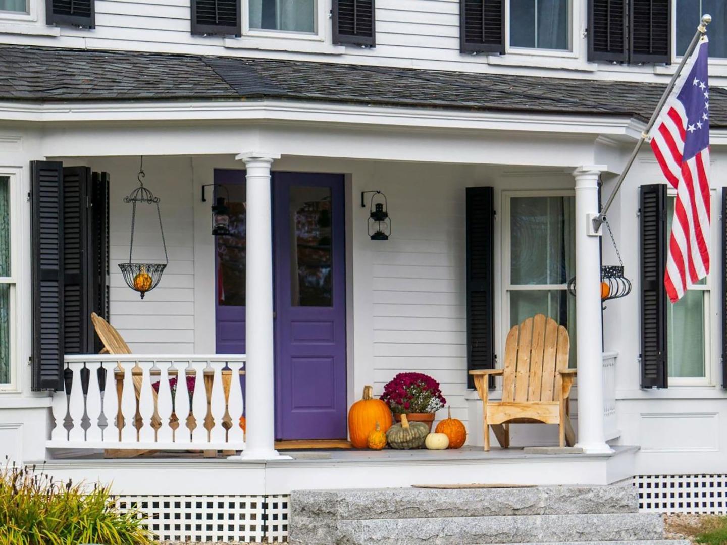 White house porch with purple door, pumpkins, wooden chair, and American flag.