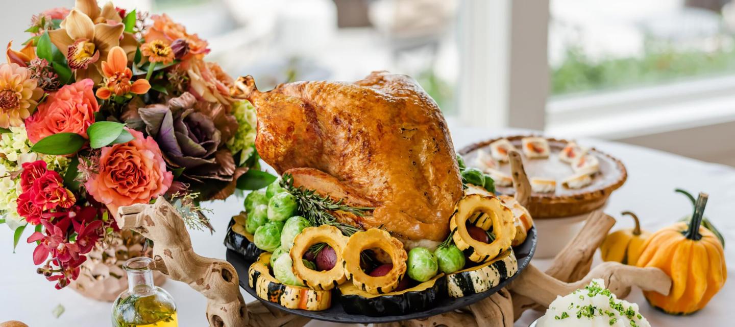 Roast turkey on a platter with flowers, pie, and pumpkins on a table.